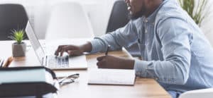 Trainee concept. African american guy taking notes while using laptop on his first day of internship in office, panorama